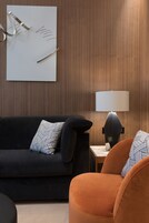 Uma sala de estar moderna com um sofá escuro, uma poltrona laranja, uma mesa lateral com abajur e uma obra de arte geométrica na parede.