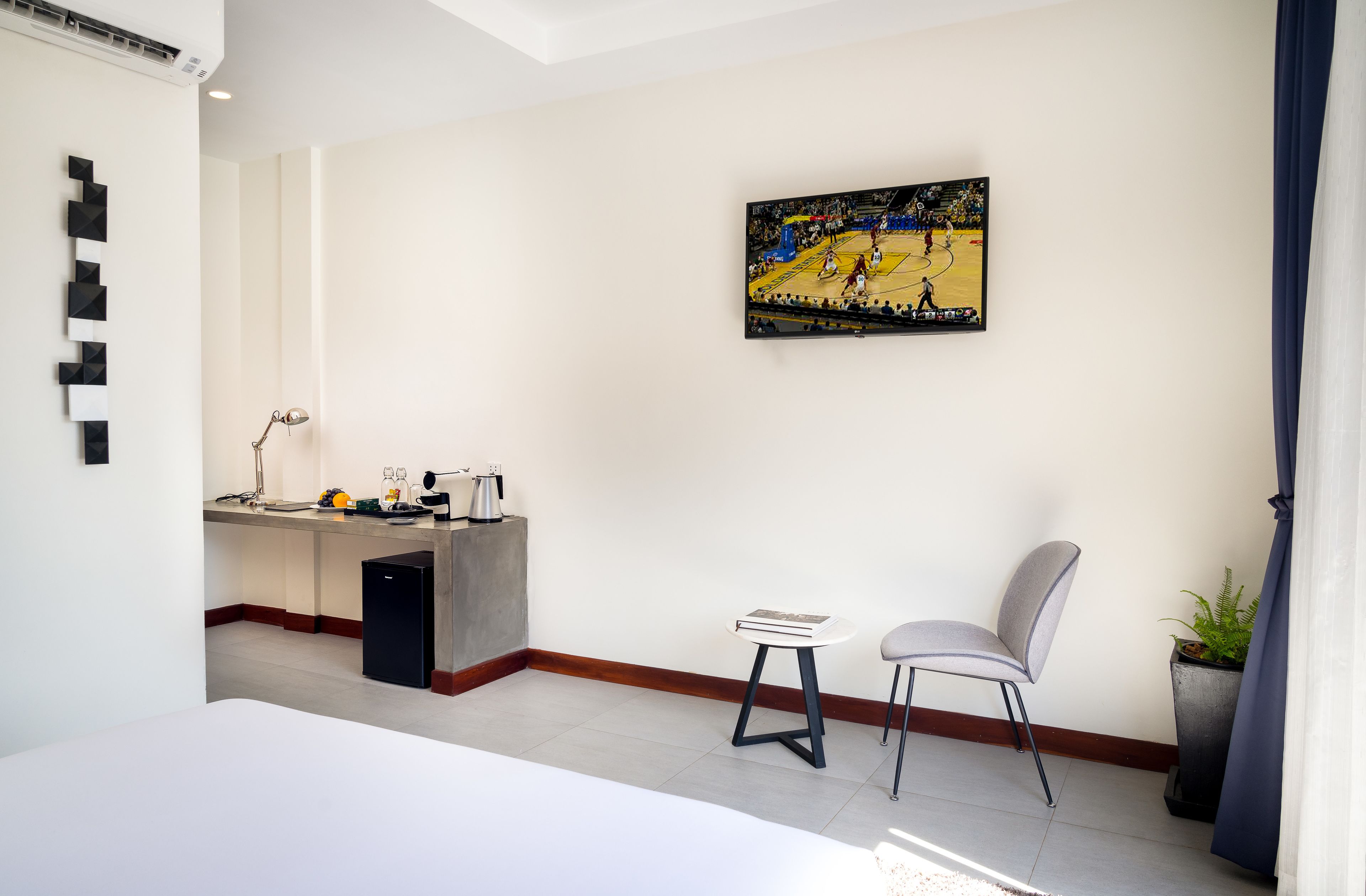 A hotel room with a bed, a desk with a lamp, a chair, a small table, a potted plant, and a sports-themed artwork on the wall.