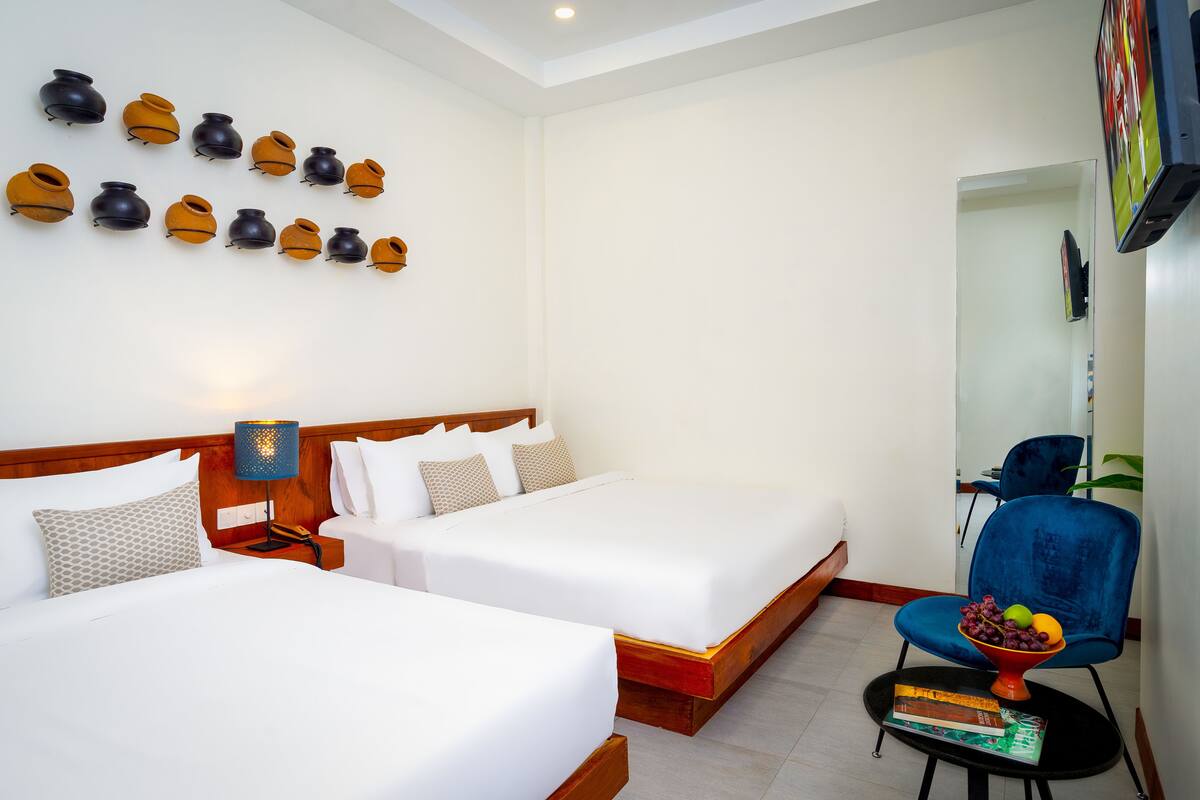 A hotel room with two beds, a blue chair, a table with a fruit bowl, and wall-mounted decorative vases.