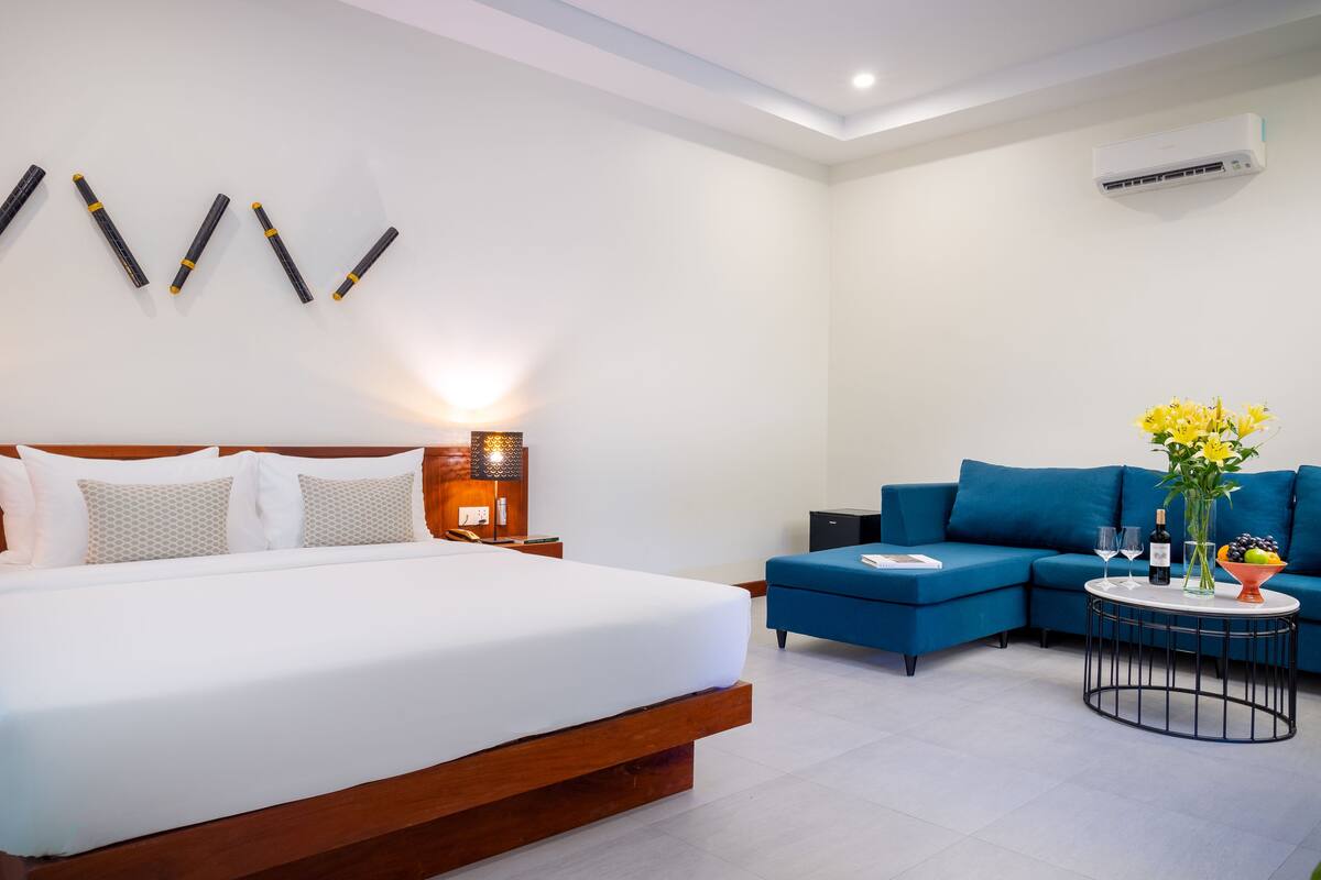 A modern hotel room with a large bed, a blue sofa, a small round table with a vase of flowers, and a wall-mounted air conditioner.
