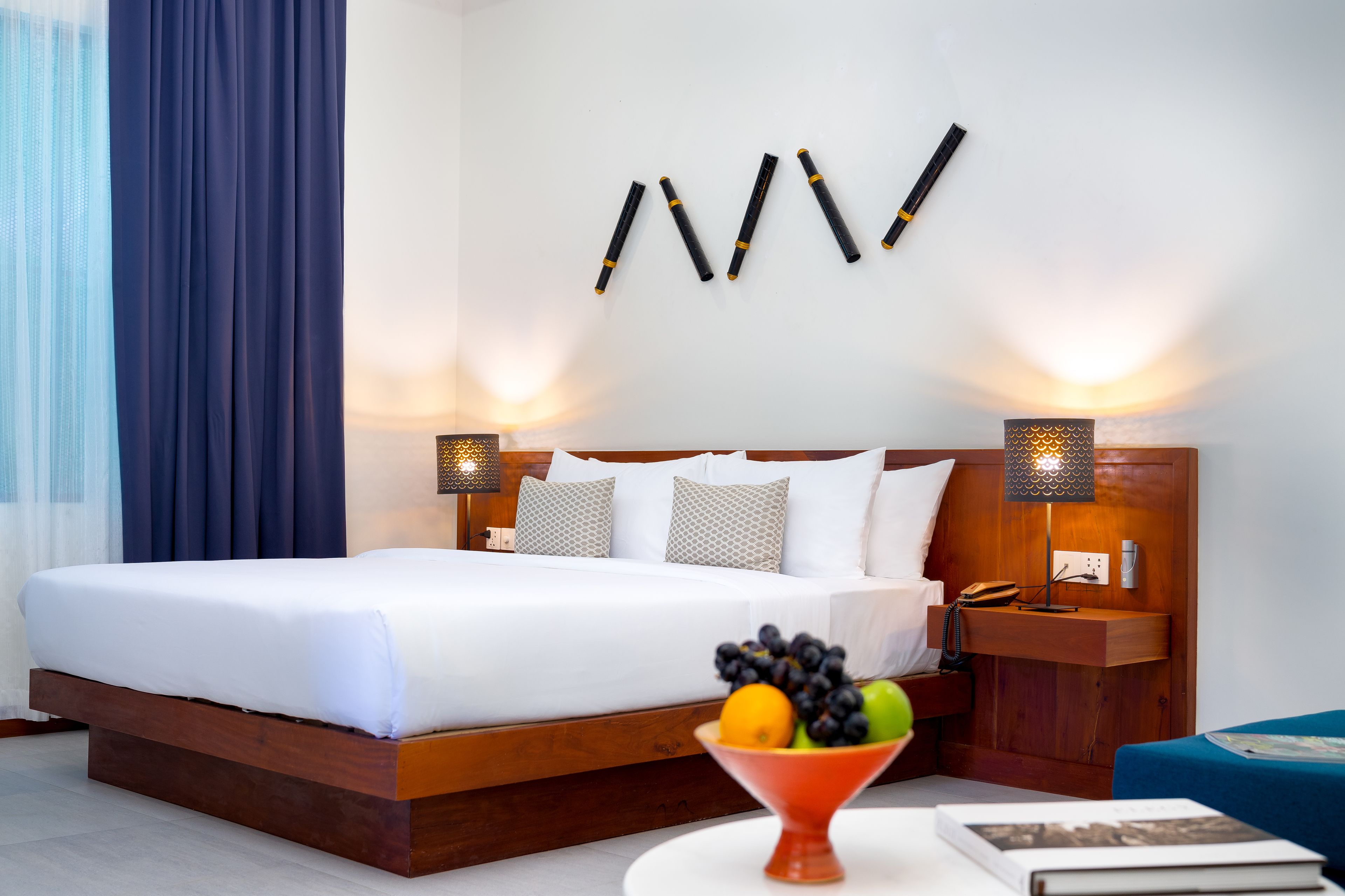 A hotel room with a large bed, bedside lamps, a fruit bowl on a side table, and a wooden nightstand with a phone.