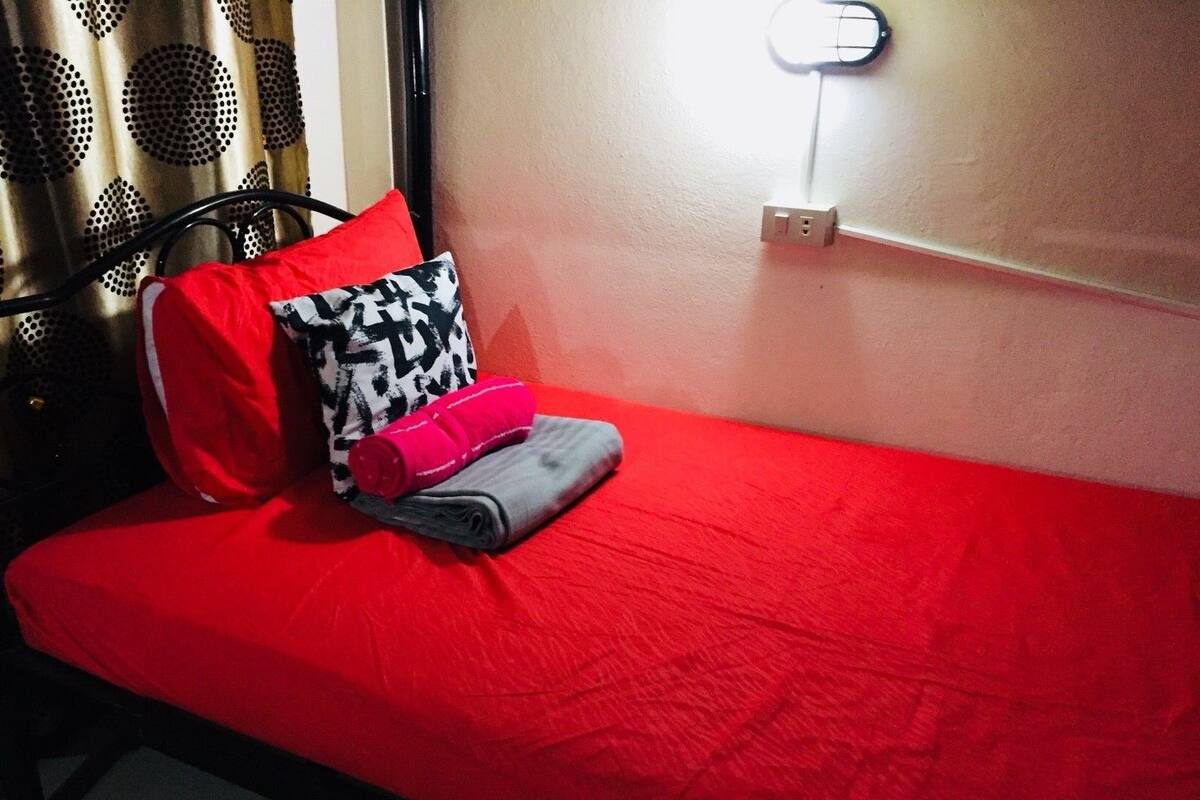 A single bed with a red bedspread, a black and white patterned pillow, a red pillow, and a folded towel.