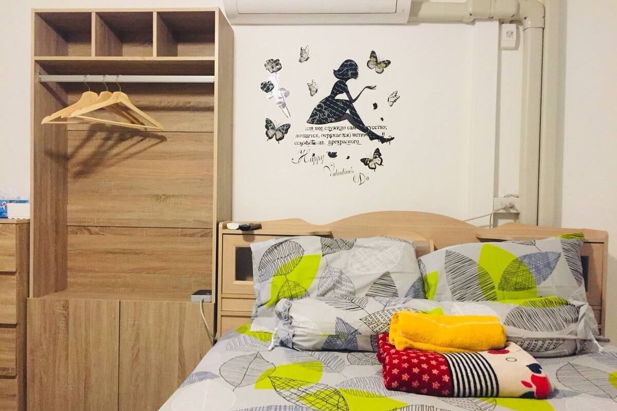 A bedroom with a wooden wardrobe, a bed with patterned bedding, and a wall decal with a silhouette of a woman and butterflies.