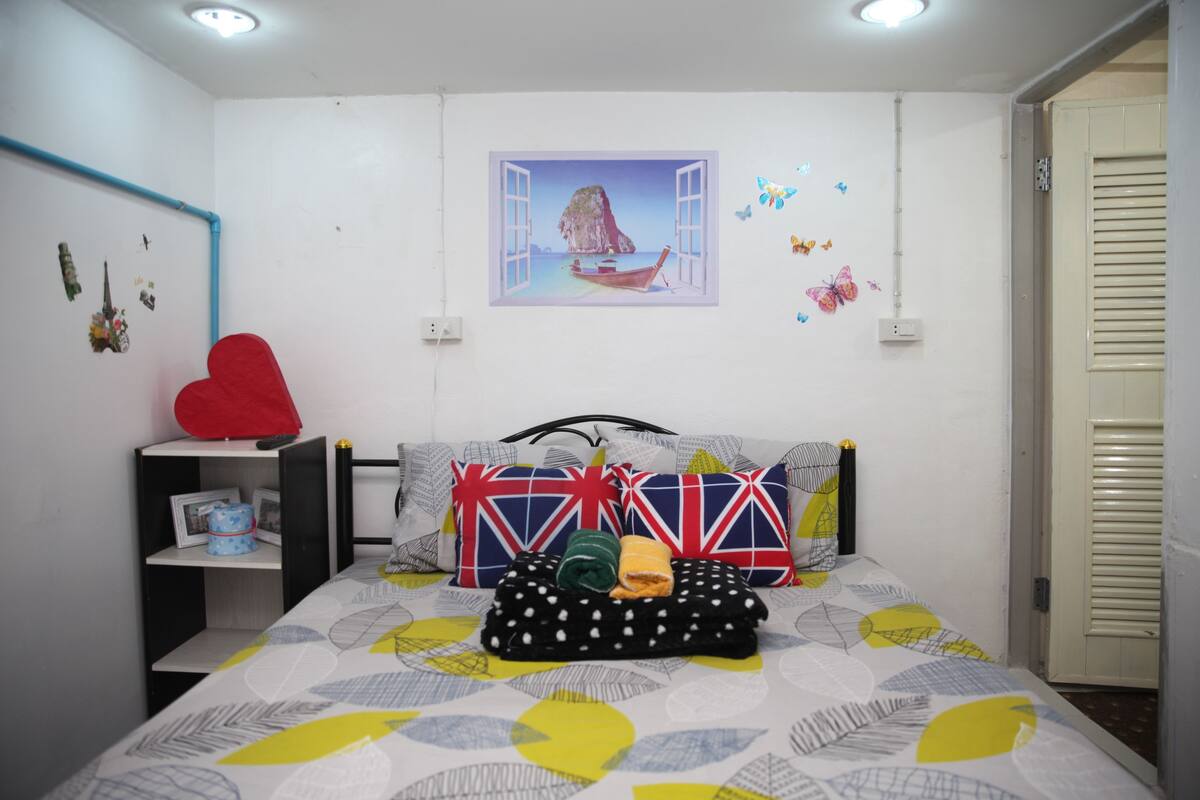 A bedroom with a bed, pillows, a shelf with items, a wall with a window view, and a door.