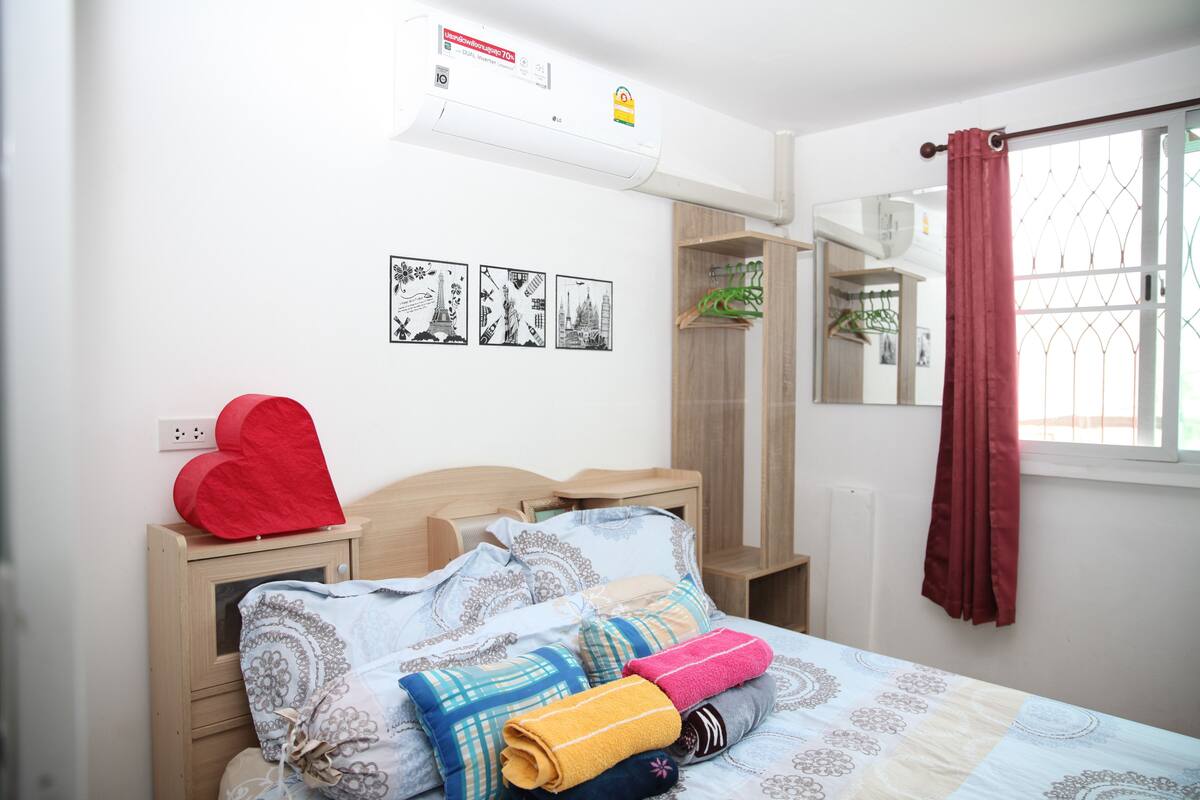 A neatly made bed with patterned bedding, a red heart-shaped decoration, and a wall-mounted air conditioner.