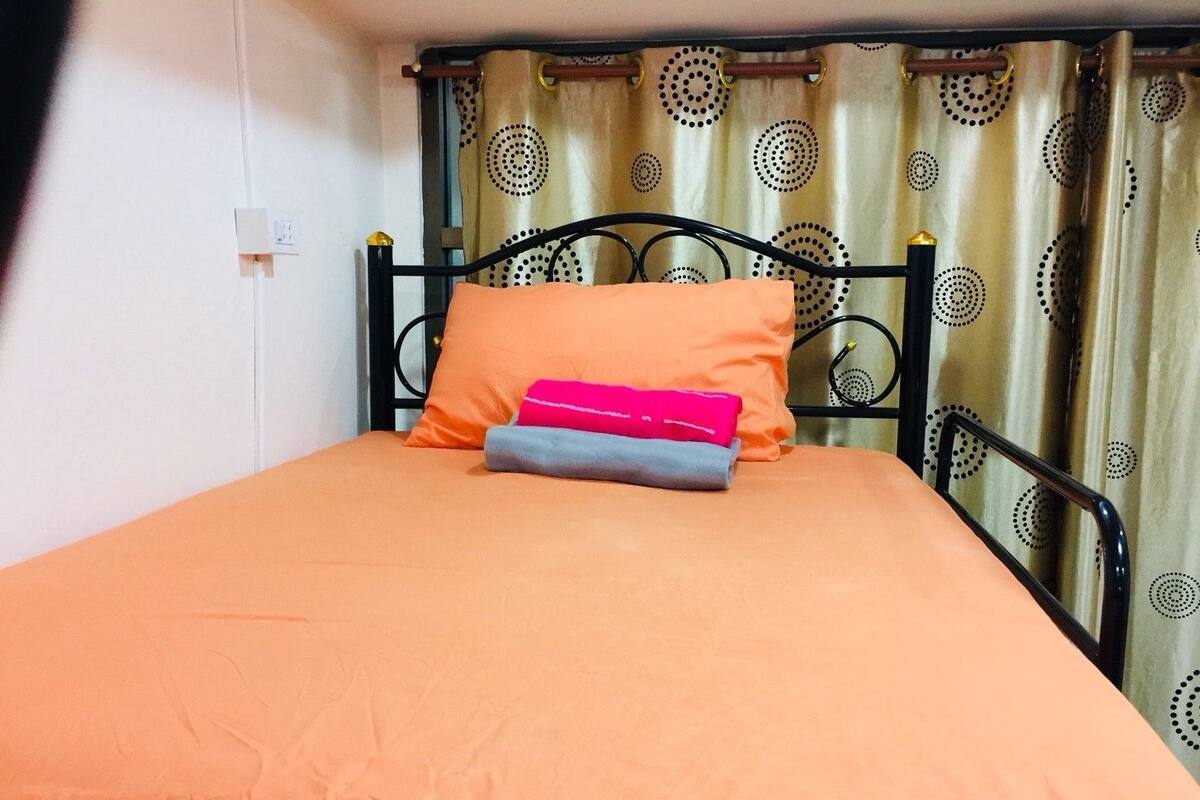 A single bed with an orange mattress, a black metal frame, and a patterned curtain in the background.
