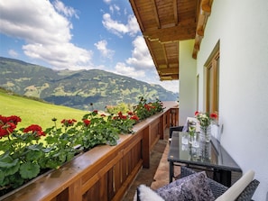 A balcony with a wooden railing, a glass table with chairs, and a view of green hills and mountains. - Apart Magdalena (Hainzenberg)