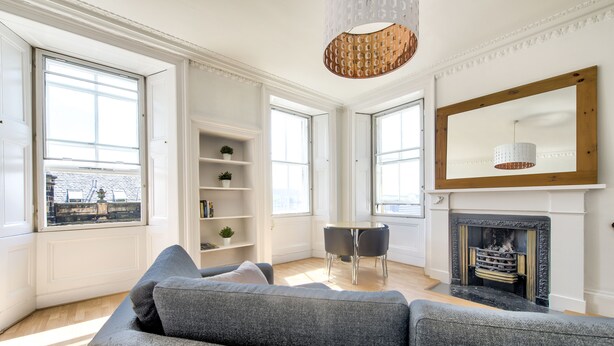 Een moderne woonkamer met een grijze bank, een eettafel met stoelen, een open haard en grote ramen.