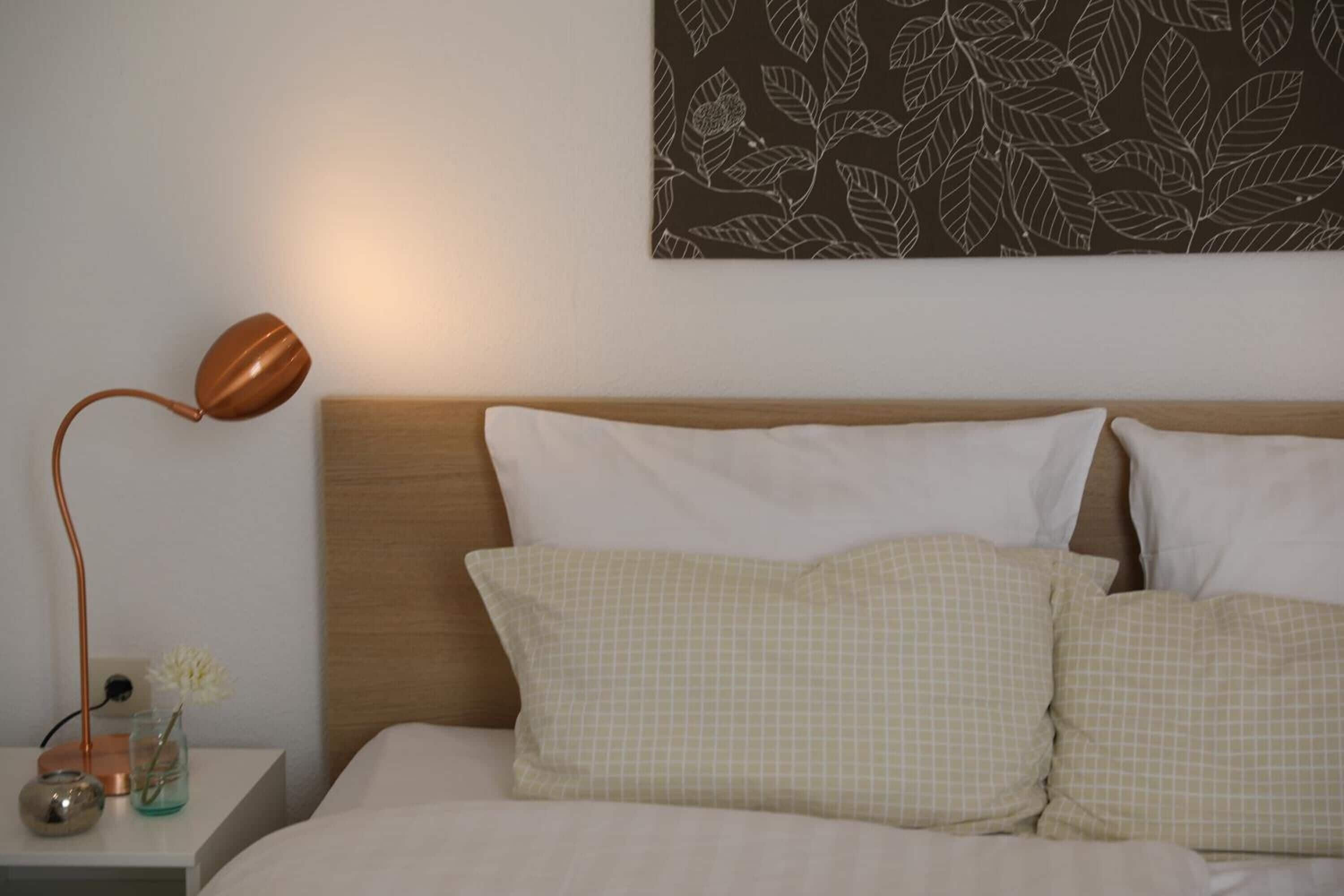 Una cama bien hecha con dos almohadas y una mesita de noche con una lámpara y un pequeño florero con flores.