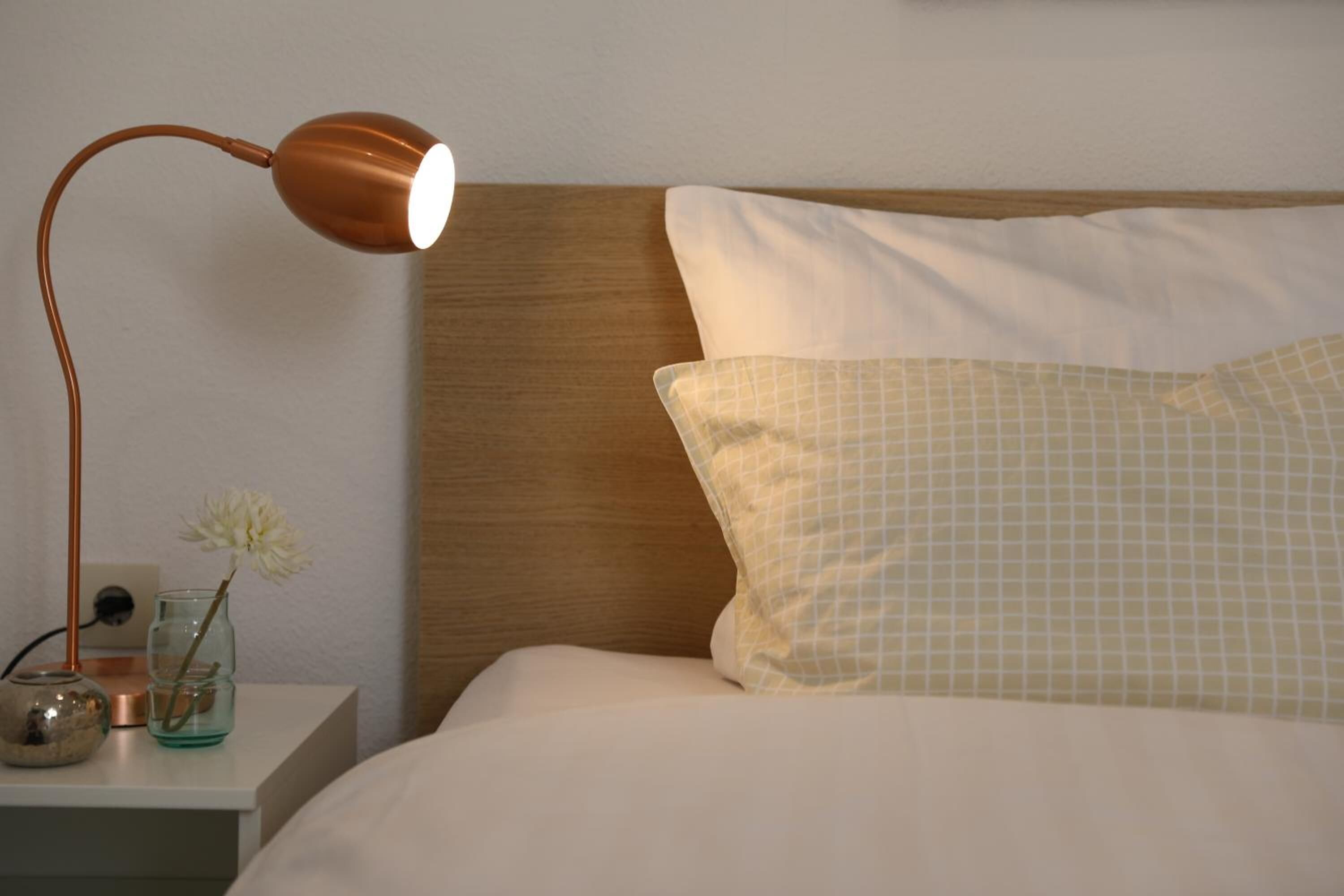 A neatly made bed with a wooden headboard, a white pillow, and a checkered pillow. A copper-colored lamp is on a bedside table next to a vase with a flower.