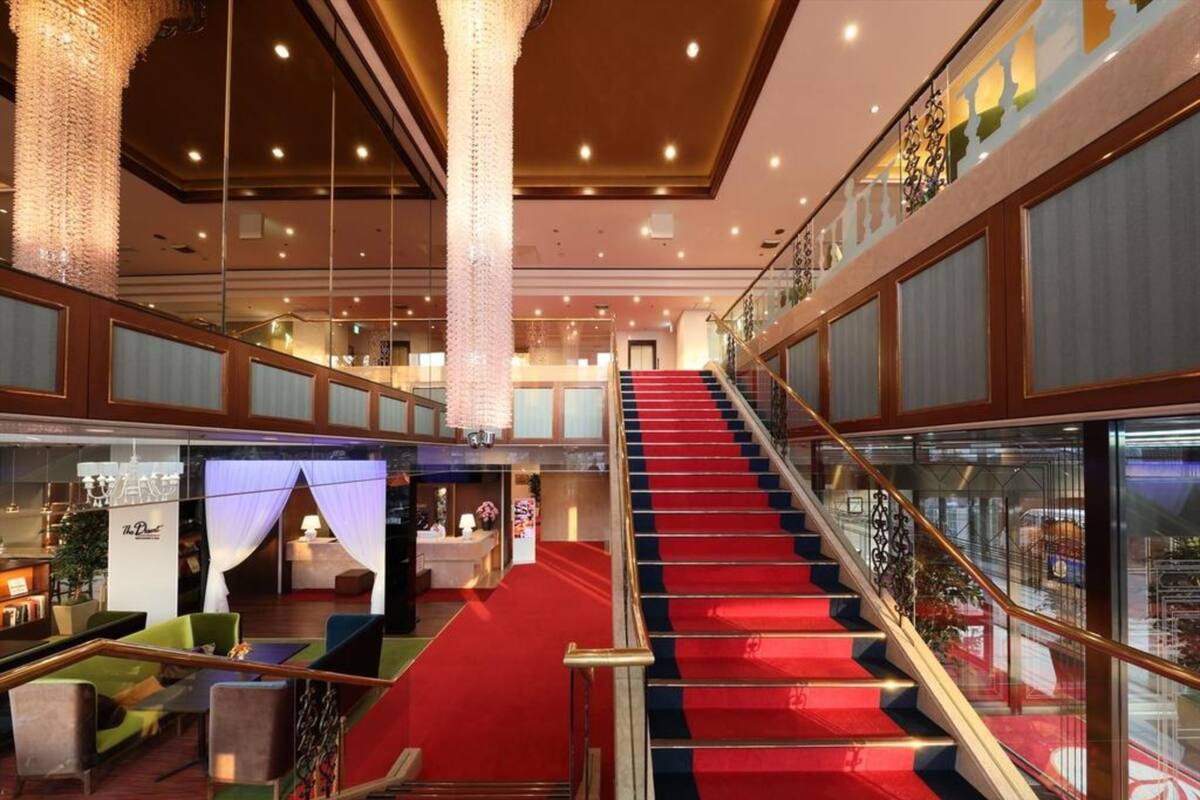 A hotel lobby with a red carpeted staircase, a reception desk, and a seating area with green sofas.