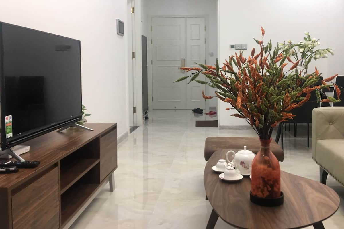 A living room with a flat-screen TV on a wooden stand, a coffee table with a vase of dried plants, a sofa, and a dining table with a teapot and cups.