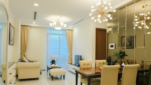 A modern living room with a sofa, ottoman, dining table, and chairs. There is a television, a plant, and a decorative light fixture. - Jack Hai Vinhome Apartment (Ho Chi Minh City)