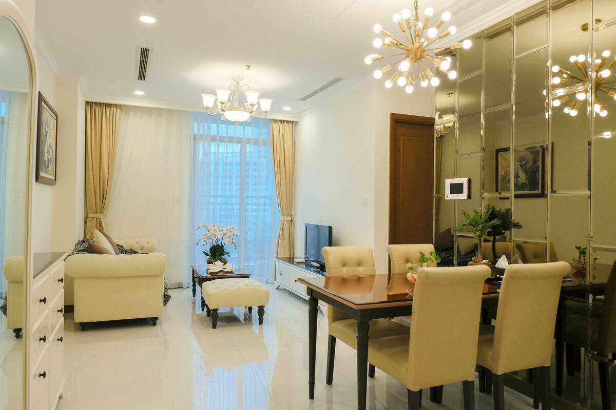 A modern living room with a sofa, ottoman, dining table, and chairs. There is a television, a plant, and a decorative light fixture.