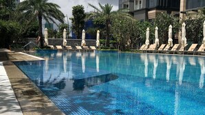 A swimming pool with lounge chairs and umbrellas, surrounded by palm trees and a stone wall. - Jack Hai Vinhome Apartment (Ho Chi Minh City)