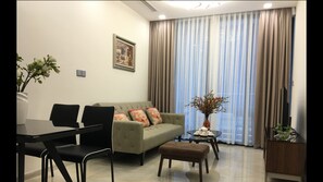 Ein modernes Wohnzimmer mit einer Couch, zwei Sesseln, einem kleinen Tisch und einem Esstisch. Ein Fenster mit Vorhängen, eine Vase mit Blumen und ein gerahmtes Bild sind an der Wand zu sehen.