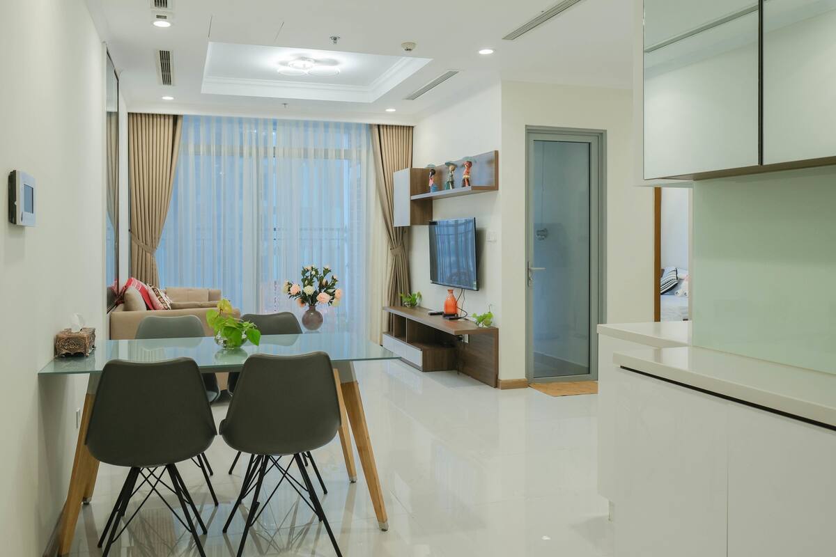 A modern living room with a glass dining table, black chairs, a sofa, and a TV on a wooden stand.