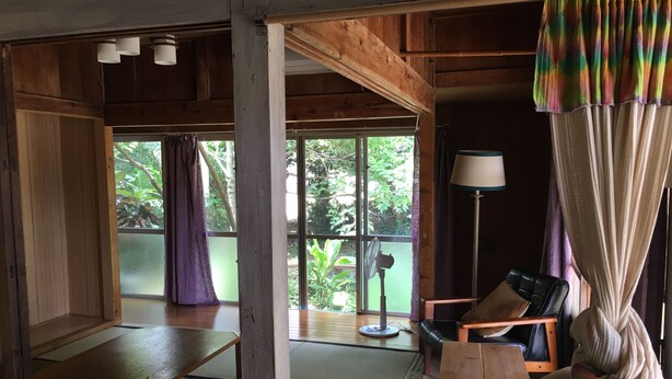 Ein traditioneller japanischer Raum mit Tatami-Böden, einem niedrigen Holztisch, einem gepolsterten Sessel, einem Standventilator und Blick auf Grünflächen.