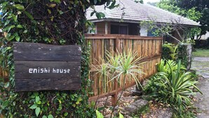 Ein Holzschild mit der Aufschrift 'enishi house', im Hintergrund ein Bambuszaun und tropische Pflanzen.
