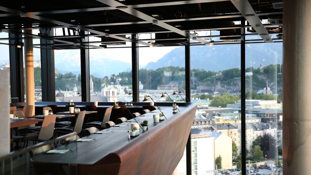 Un restaurante moderno con una larga mesa de madera y sillas, amplias ventanas que ofrecen vista a la ciudad y montañas al fondo.