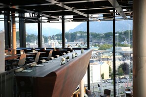 Un restaurante moderno con una larga mesa de madera y sillas, amplias ventanas que ofrecen vista a la ciudad y montañas al fondo.