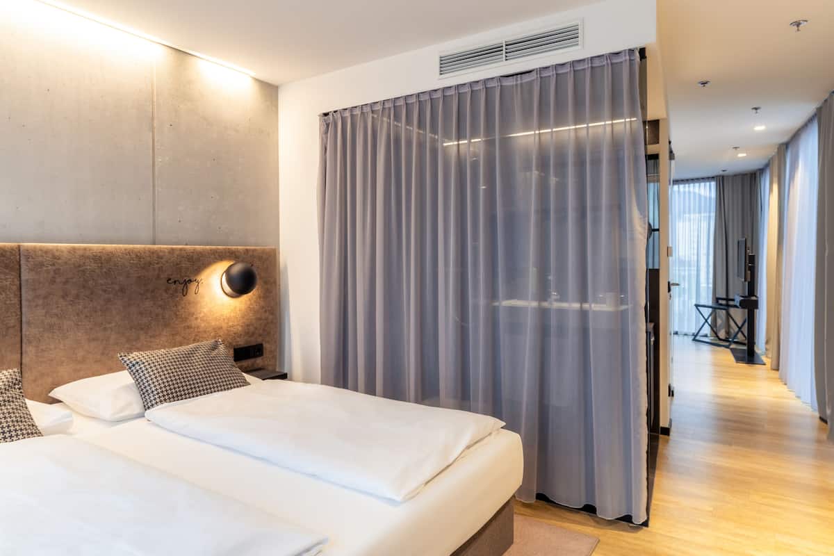 Ein modernes Hotelzimmer mit einem großen Bett, einem Kopfteil mit Leuchte und Blick auf einen Flur mit einer Sitzecke.