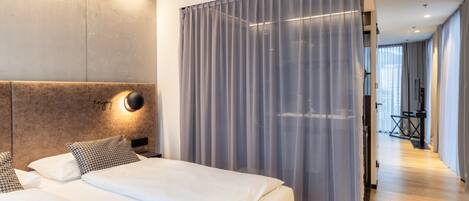 Una habitación de hotel moderna con una cama grande, un cabecero con una lámpara y vistas a un pasillo con zona de estar.