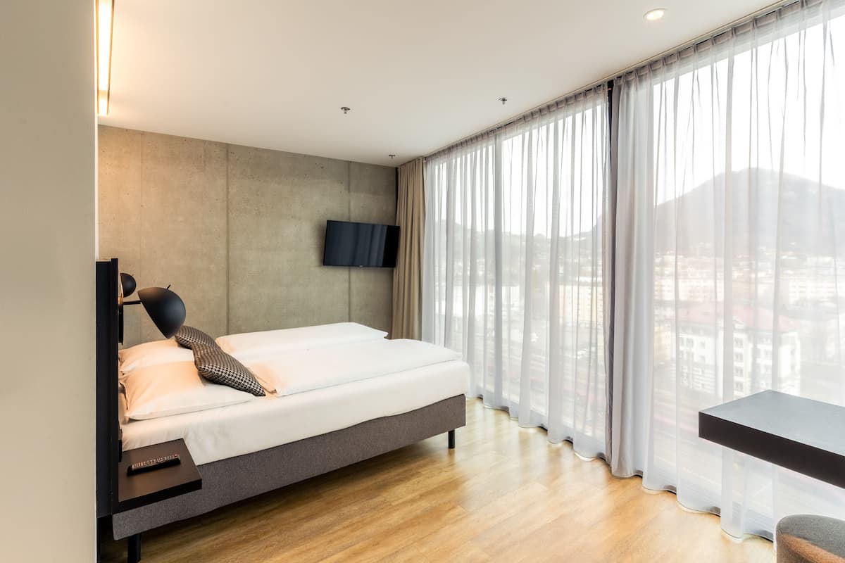 Ein modernes Schlafzimmer mit einem Bett, einem an der Wand befestigten Fernseher, einem Schreibtisch und großen Fenstern mit durchscheinenden Vorhängen.