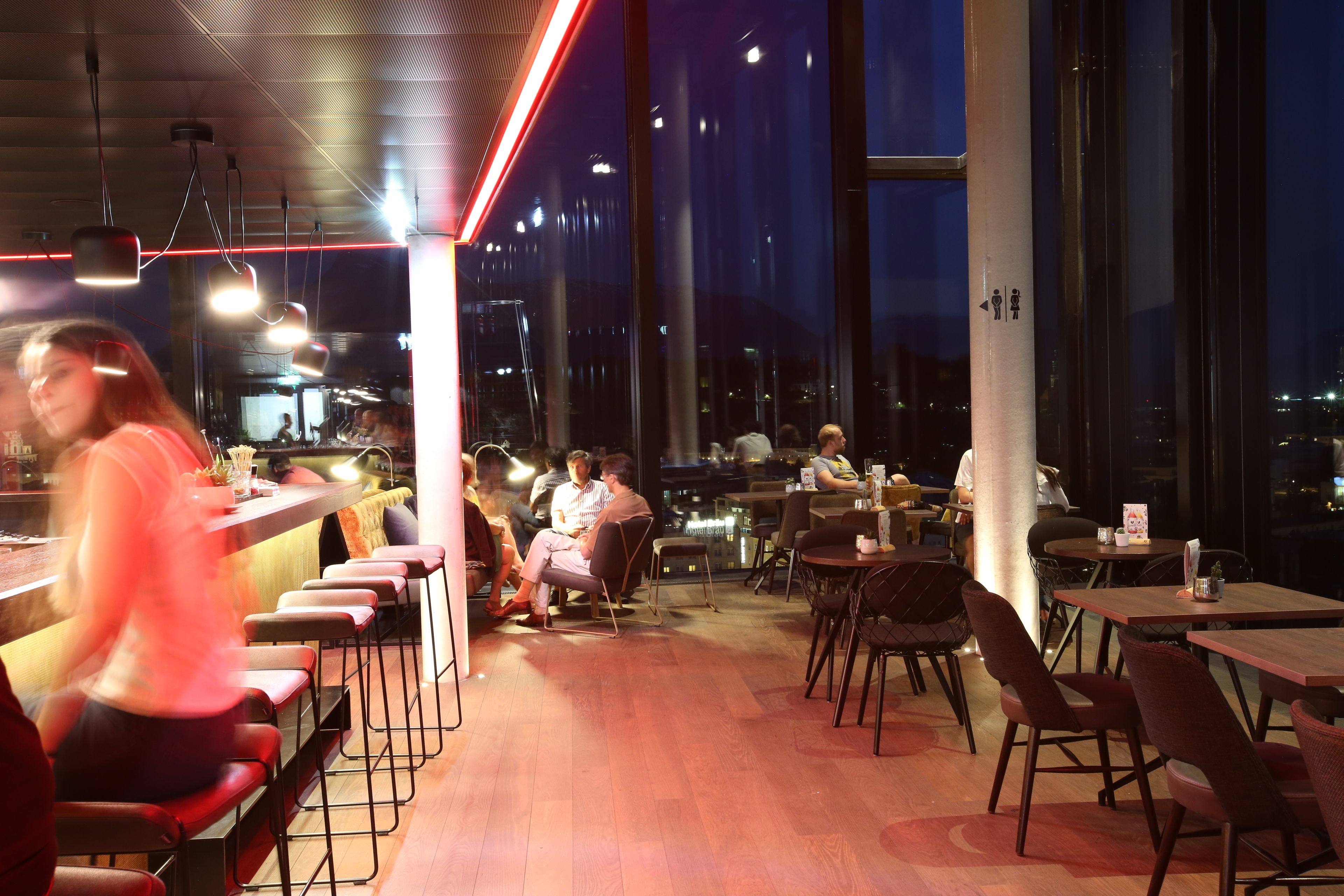Un restaurante moderno con techos altos, ventanas amplias y una variedad de opciones de asientos, incluyendo taburetes de bar y mesas.