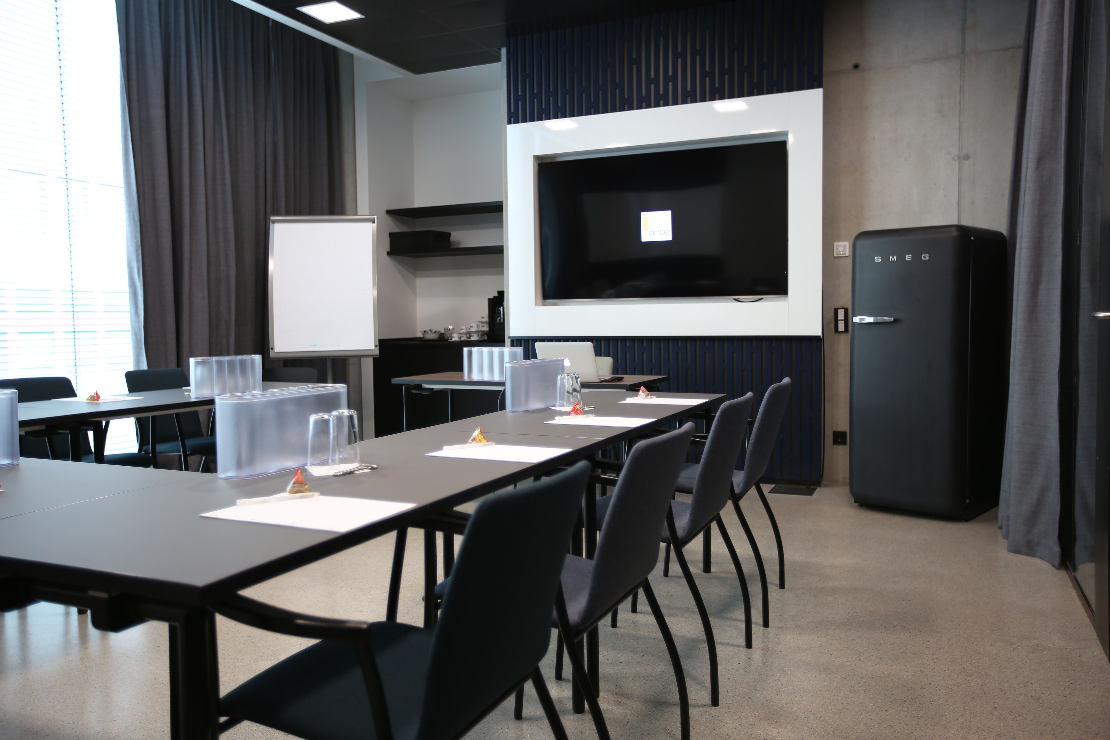 A conference room with a long table, chairs, a projector screen, a whiteboard, and a refrigerator.