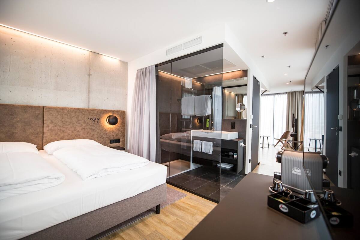 Ein modernes Hotelzimmer mit einem großen Bett, einem in Glas eingefassten Badezimmer und einem Schreibtisch mit Kaffeemaschine.