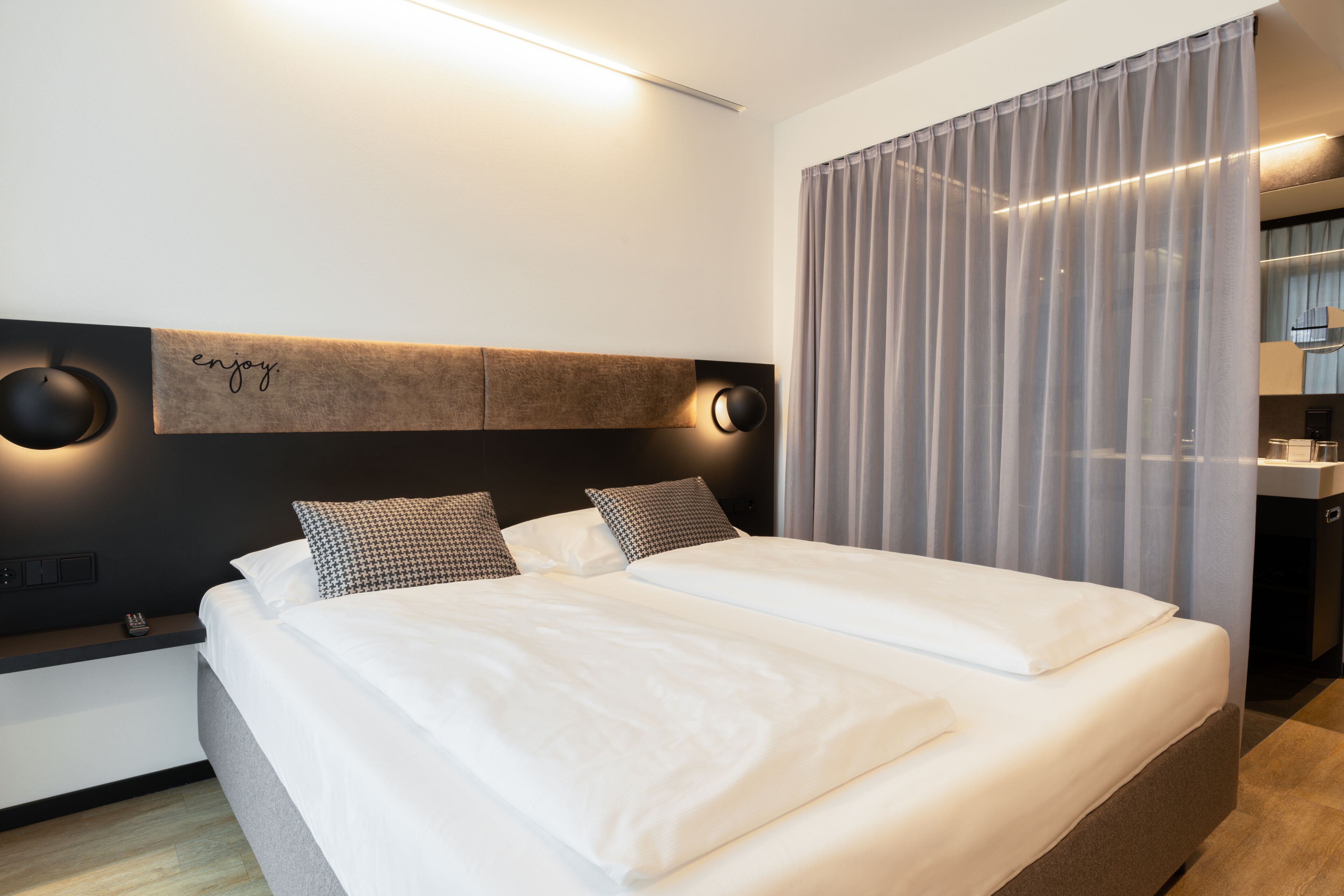 A modern hotel room with a large bed, a headboard with ambient lighting, a bedside table, and a view of a cityscape through sheer curtains.