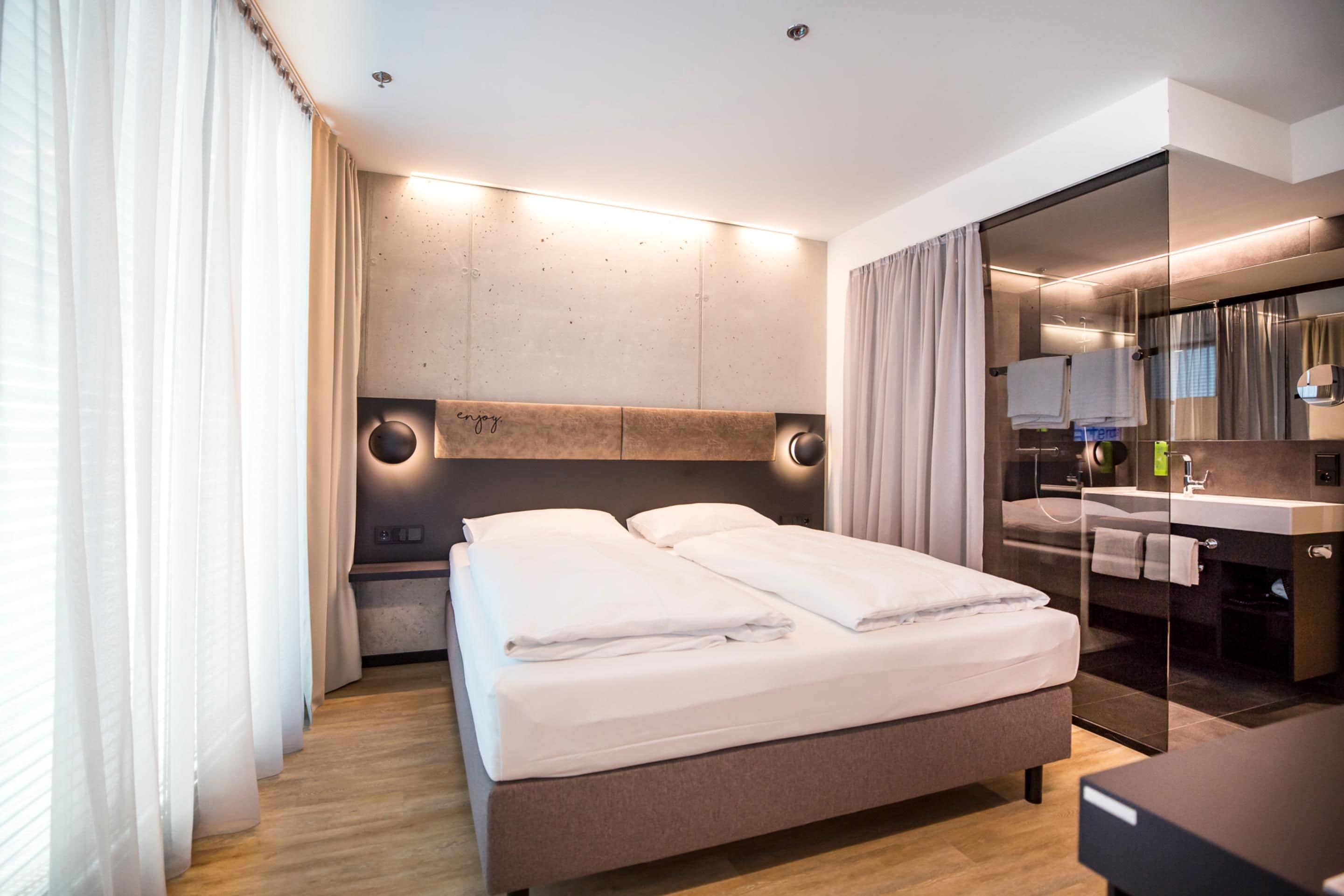 Ein modernes Hotelzimmer mit einem großen Bett, einer Duschkabine aus Glas und einem separaten Badezimmer mit Waschtisch.