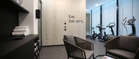 A modern fitness center with exercise equipment, a reception area with a counter and towels, and a motivational wall with the phrase "be fit. be art."