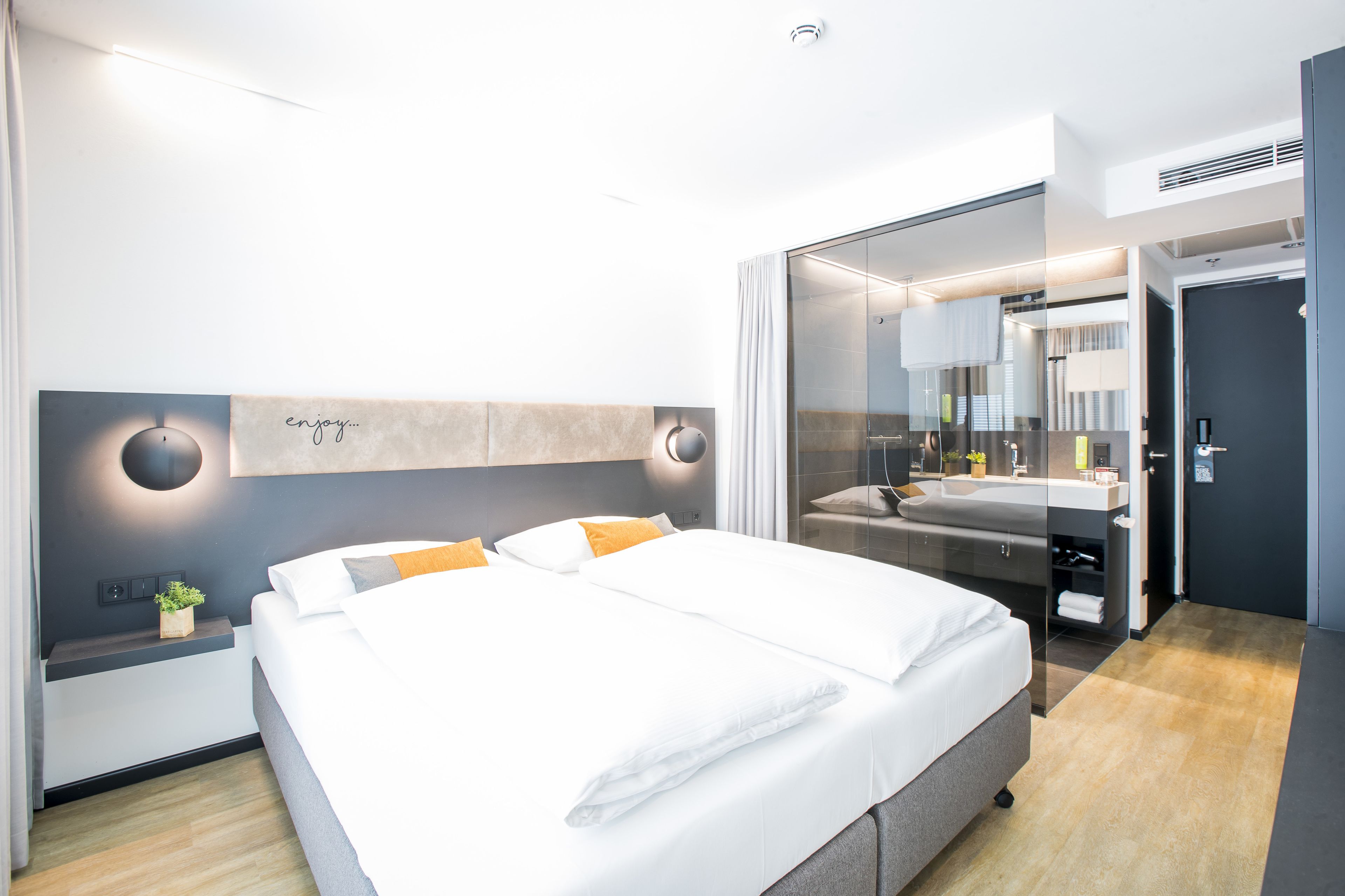 A modern hotel room with a large bed, bedside tables, a mirror, and a bathroom with a sink and shower.