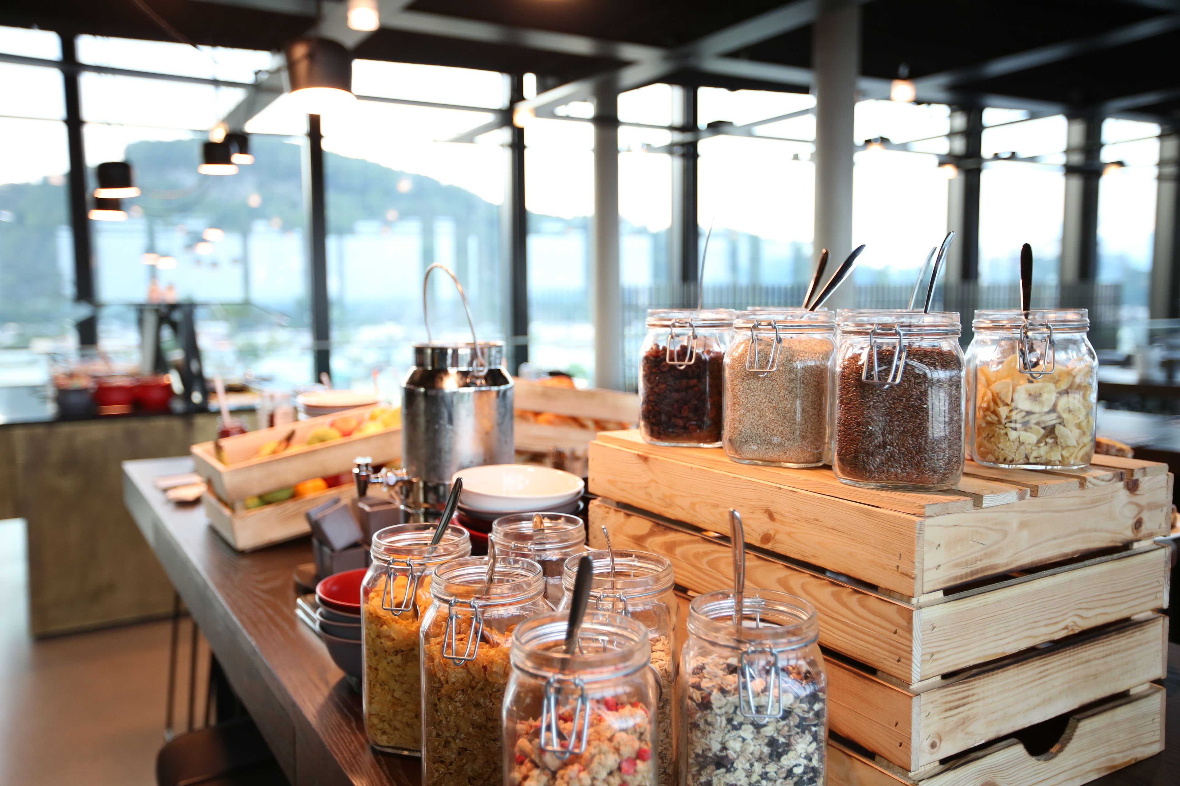 Montaje de bufé con diversos alimentos en frascos y recipientes, una cafetera y vistas de la ciudad a través de amplios ventanales.
