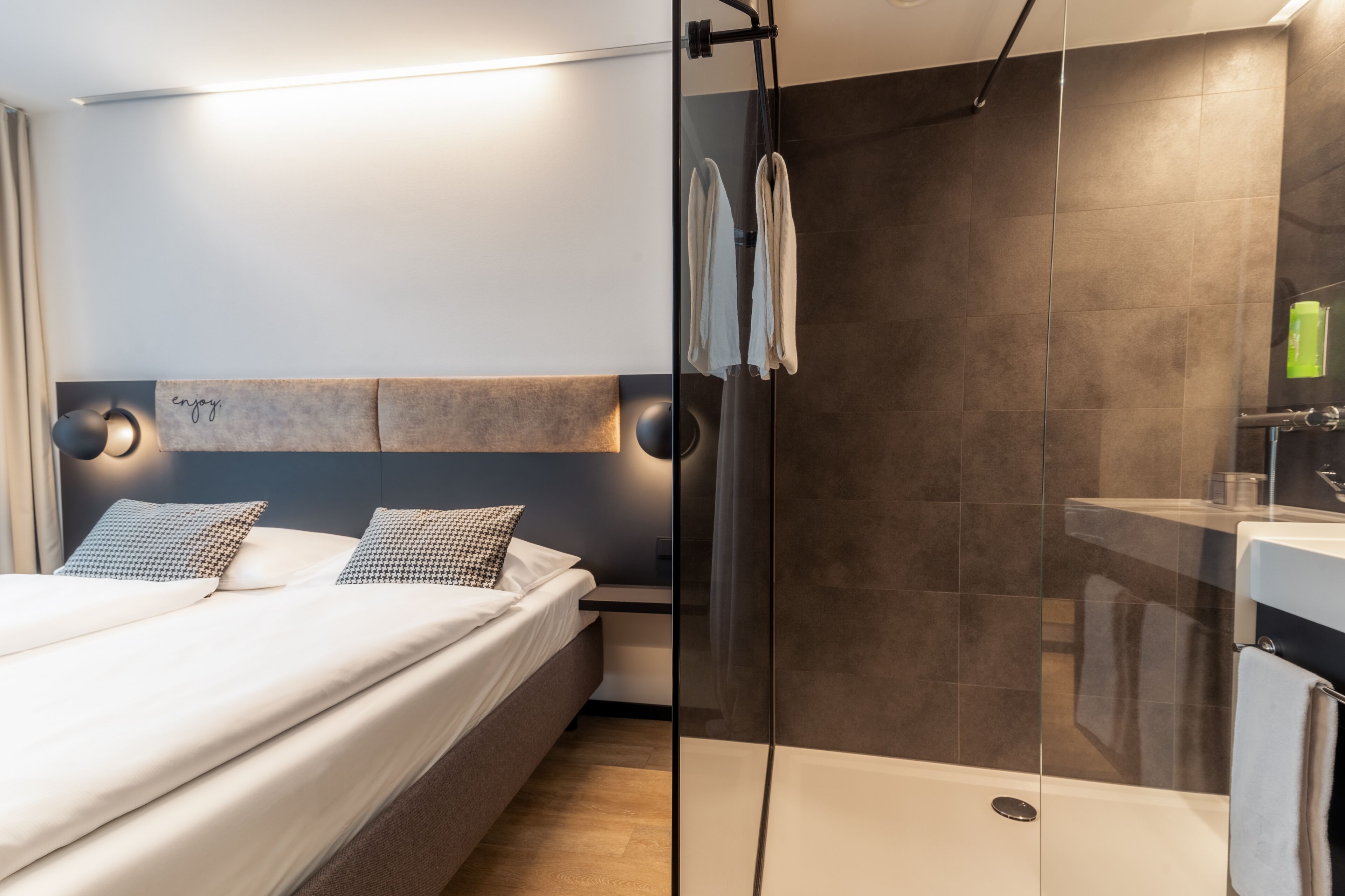 A modern hotel room with a bed, a glass shower enclosure, and a bathroom with a sink and mirror.