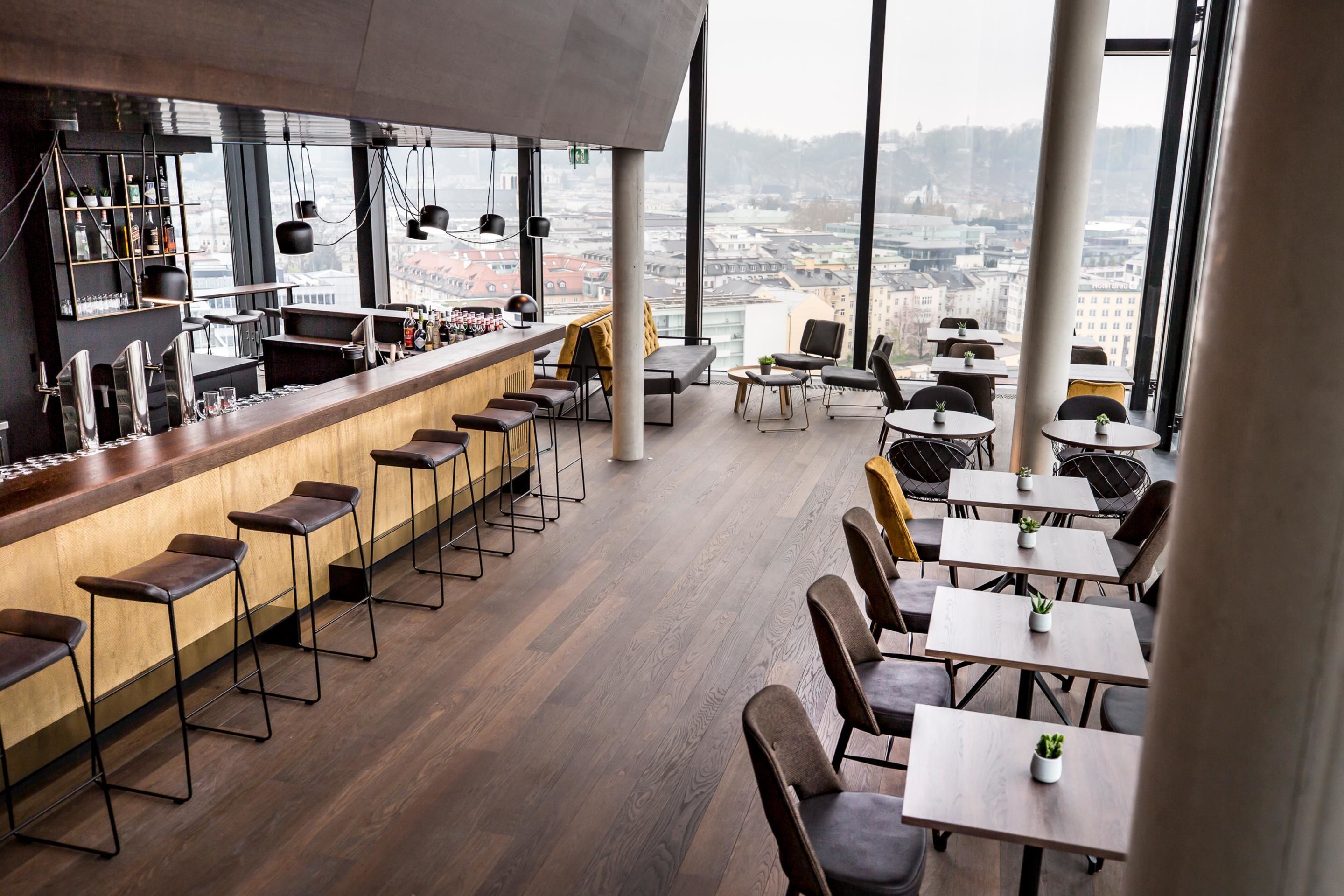 A modern bar area with a long counter, stools, and a view of the city through large windows.
