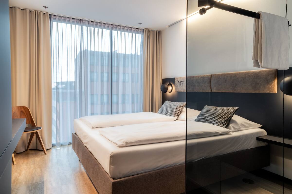 Ein modernes Hotelzimmer mit einem großen Bett, einem Schreibtisch und Blick auf die Stadt durch transparente Vorhänge.