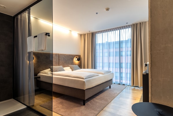 Habitación de hotel moderna con una cama grande, cabina de ducha de vidrio y vistas de la ciudad a través de amplios ventanales.