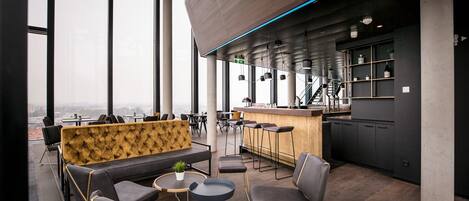 A modern lounge area with a sofa, chairs, and tables, featuring a city view through large windows.
