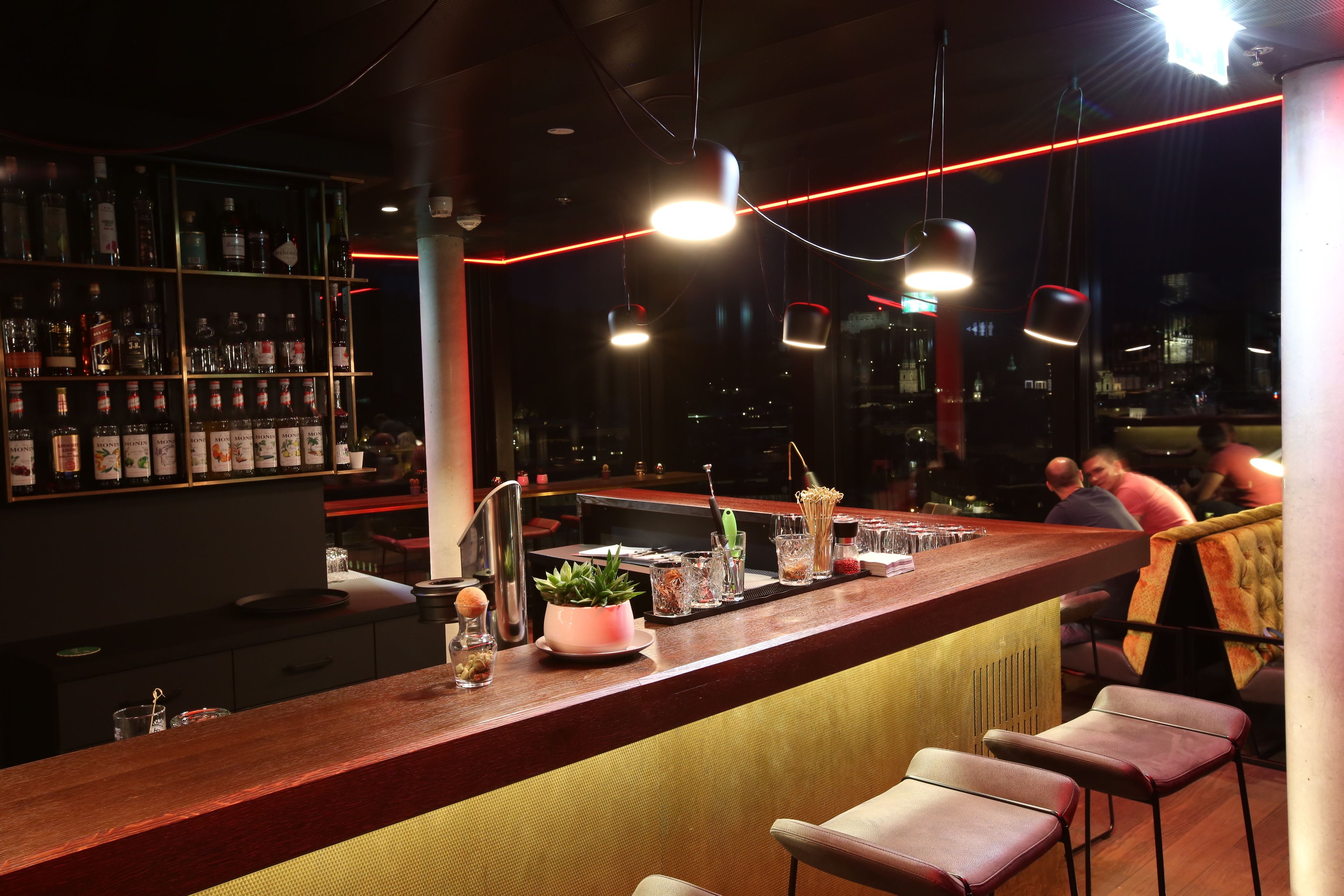 A bar area with a wooden counter, shelves with bottles, and hanging pendant lights.