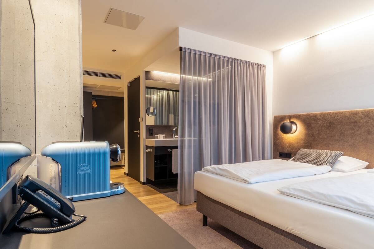 Ein Hotelzimmer mit einem Bett, einem Schreibtisch mit Telefon, einem Spiegel und einem durch eine Tür einsehbaren Badezimmer.