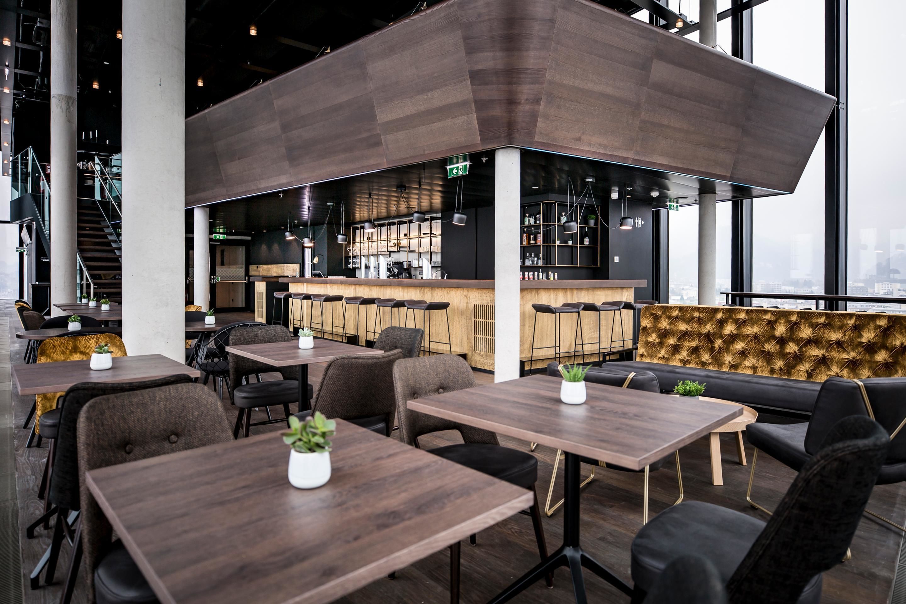 A modern restaurant with wooden tables and black leather seating, a bar area, and large windows offering a city view.