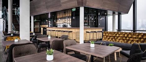 A modern restaurant with wooden tables and black leather seating, a bar area, and large windows offering a city view.
