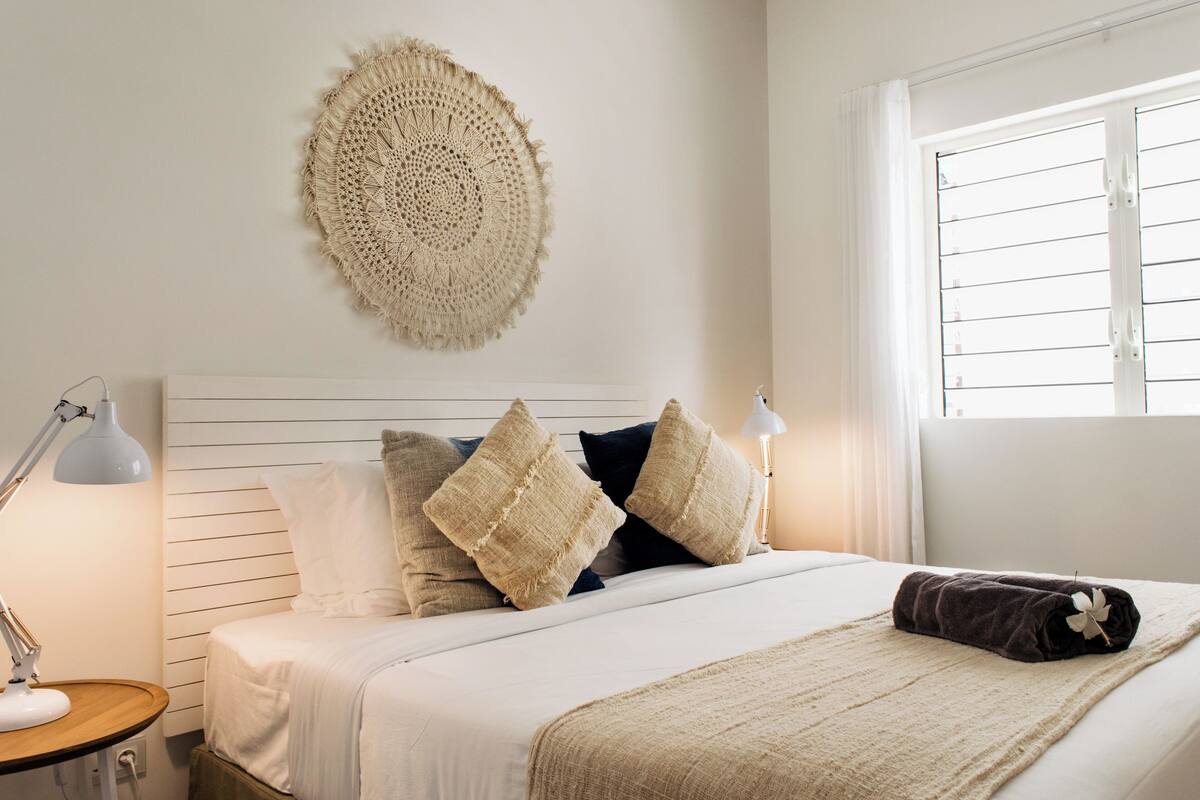 A neatly made bed with a white headboard, a woven wall hanging, and a window with blinds.