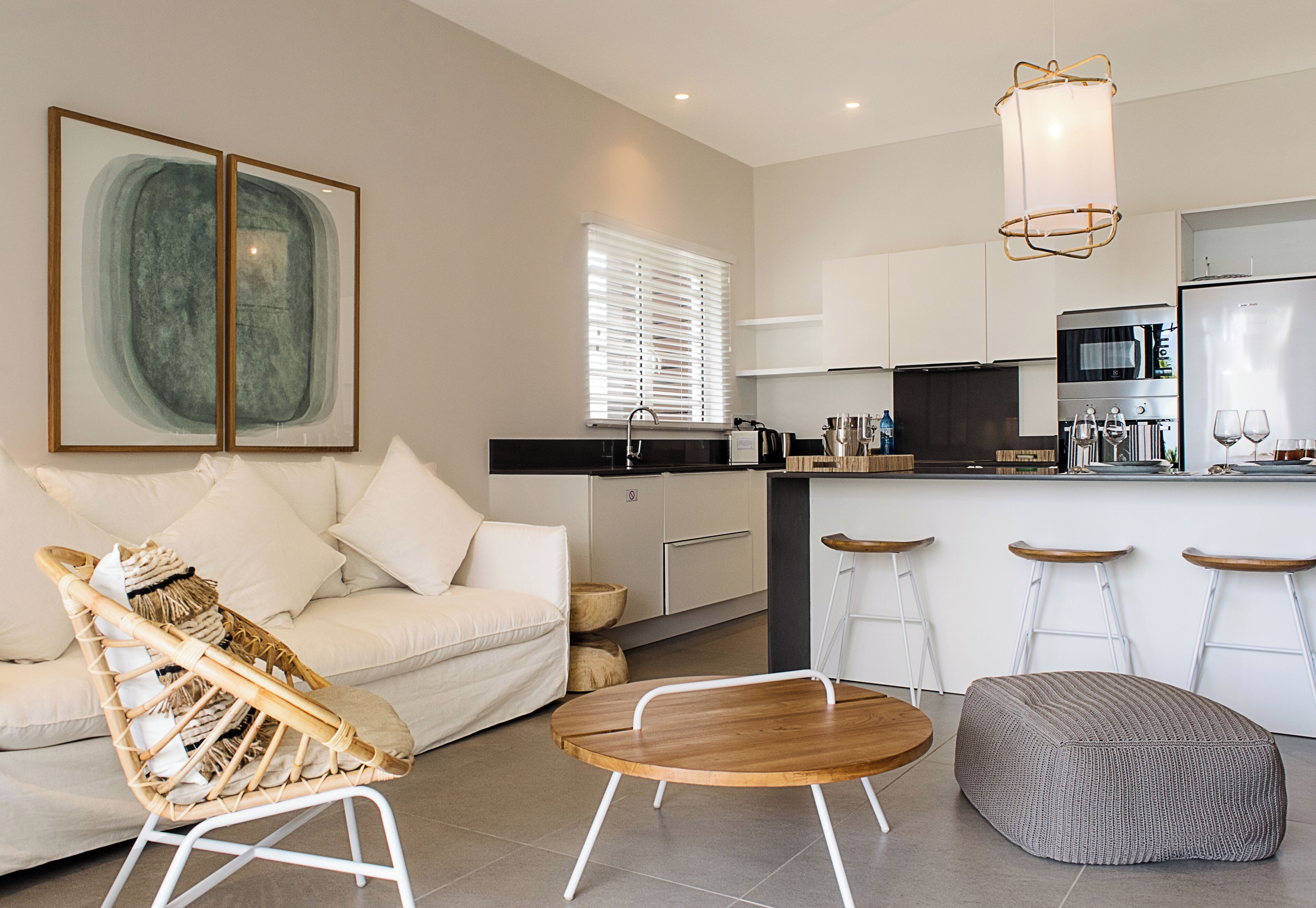 Una sala moderna con un sofá, una mesa de centro de madera, una silla de mimbre y un pouf gris. Hay una zona de cocina con un refrigerador, horno y taburetes de bar.