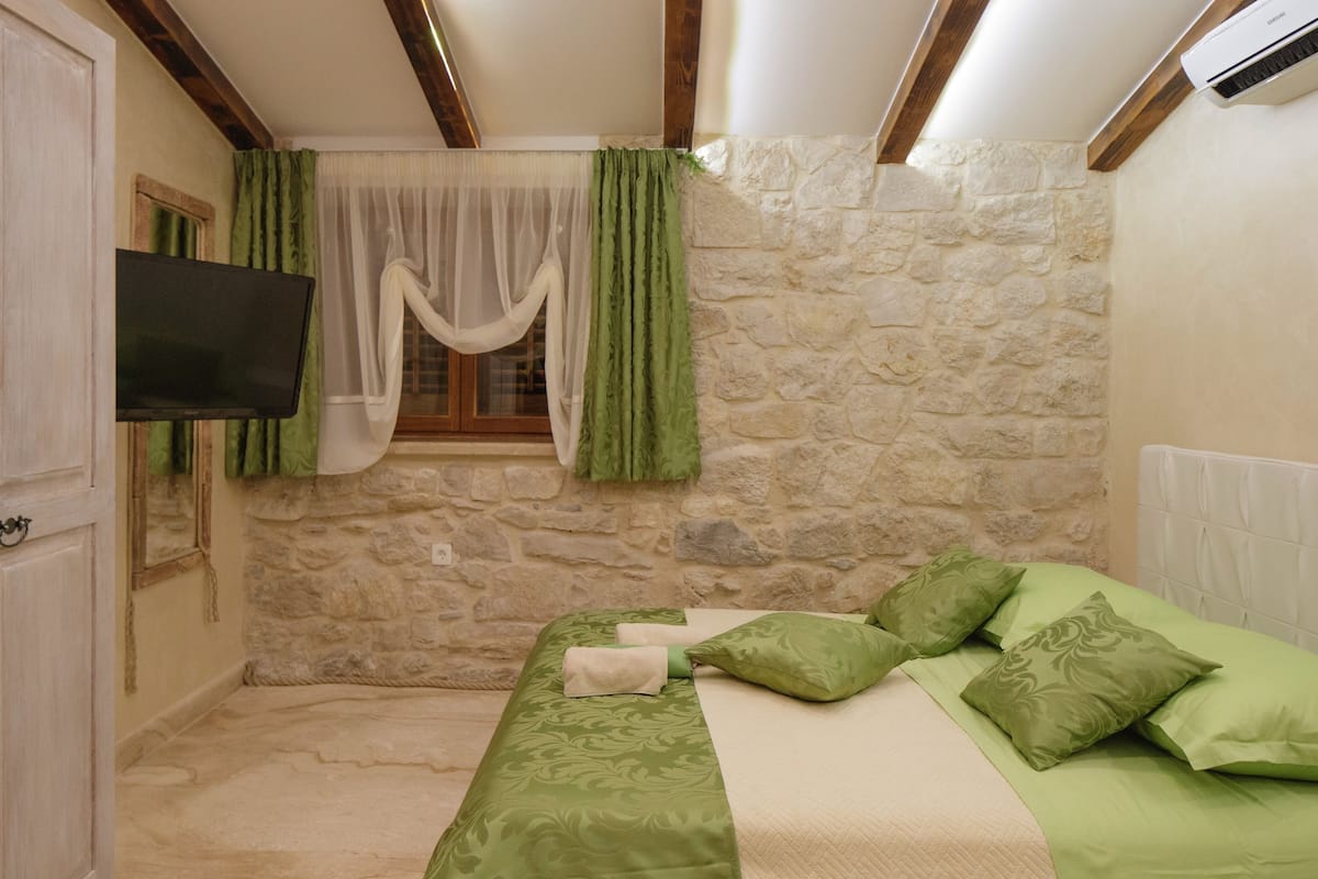 A bedroom with a stone wall, a bed with green bedding, a TV mounted on the wall, and a window with green curtains.