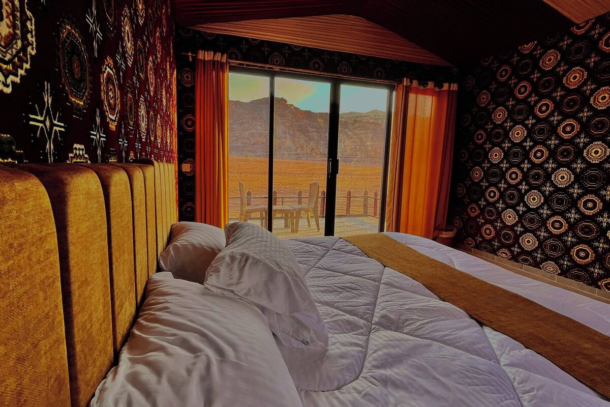 Una habitación con una cama grande, vistas a las montañas a través de un ventanal corredero, paredes con un diseño decorativo y un techo de madera.