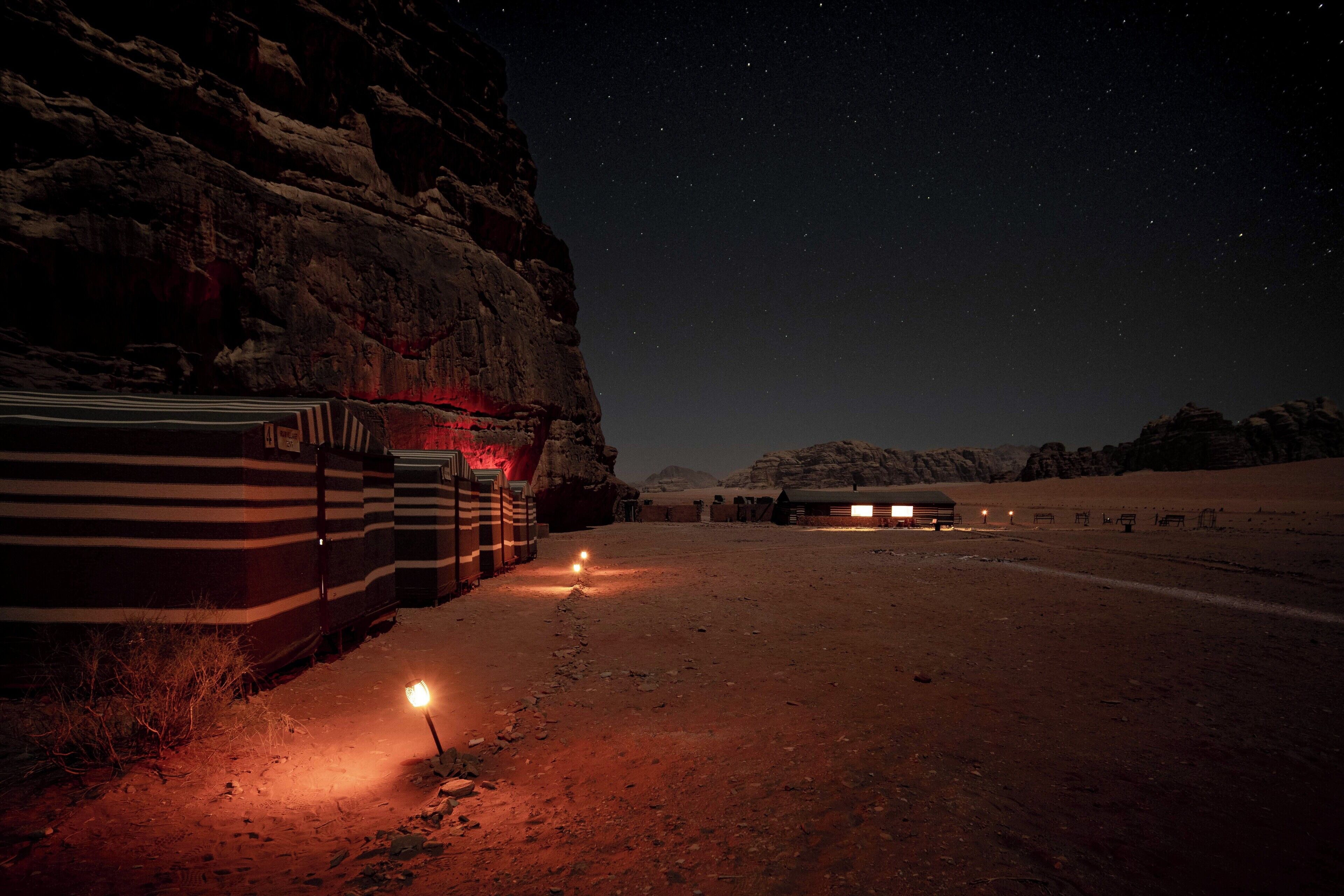A desert landscape at night with a row of striped tents and a few lit windows, suggesting a campsite or caravan park.